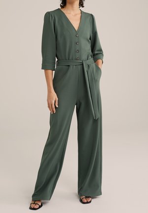 Kobieta ubrana w oliwkowo zielony kombinezon zapinany na guziki z rękawami 3/4 i paskiem do wiązania, ręce w kieszeniach, w połączeniu z czarnymi sandałami z odkrytymi palcami.