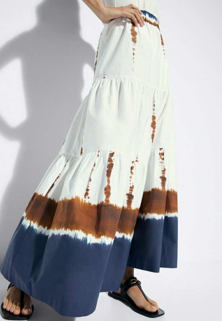 Femme portant une jupe longue à volants avec un motif tie-dye blanc, marron et bleu marine, associée à des sandales noires à lanières, debout avec la main sur la hanche.