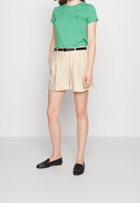 Vihreä puuvillapaita, jossa on pyöreä pääntie, beige pystysuoraraidalliset shortsit, joissa on taskut ja musta kiiltävä vyö, yhdistettynä mustiin loafer-kengiin.