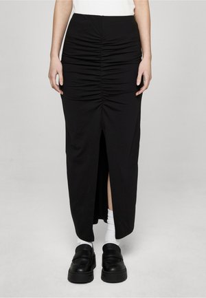 Kvinde iført en sort midi-nederdel med rynk og frontslids, hvide sokker, sorte chunky loafers og en hvid indstoppet top, stående mod en ensfarvet baggrund.