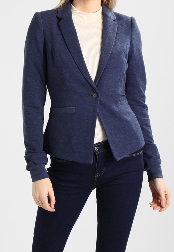 Femme portant un blazer bleu marine ajusté avec boutons à l'avant et deux poches, associé à un jean bleu foncé et un haut blanc côtelé en dessous.