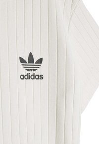 Λευκό ριγωτό ύφασμα με μαύρο λογότυπο Adidas που απεικονίζει τρεις ρίγες και το τριφύλλι στο στήθος. Κοντά μανίκια και υφή υφάσματος.