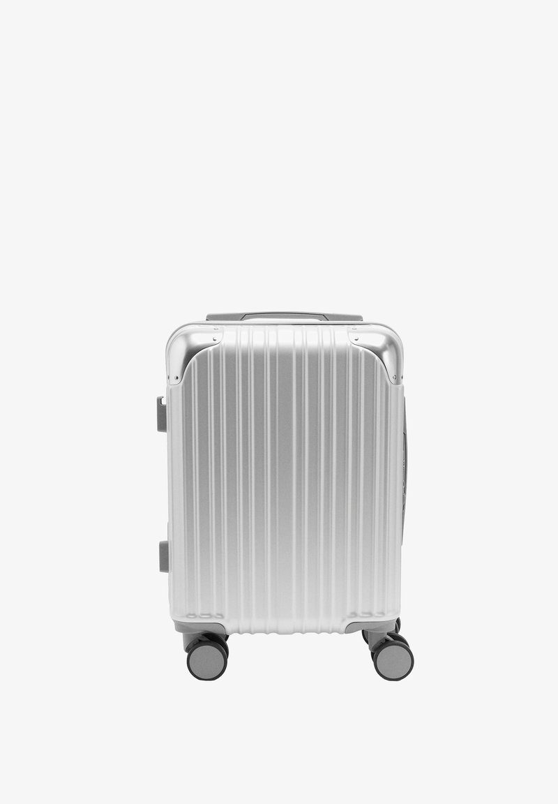 Srebrna walizka z twardą obudową, z pionowymi rowkami, wysuwanym uchwytem i czterema kółkami dla łatwości przemieszczania. Posiada zaokrąglone narożniki i solidną konstrukcję.