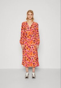 Kvinna som står mot en enkel bakgrund, iförd en långärmad, V-ringad, orange och rosa blommig midiklänning med vita sneakers.