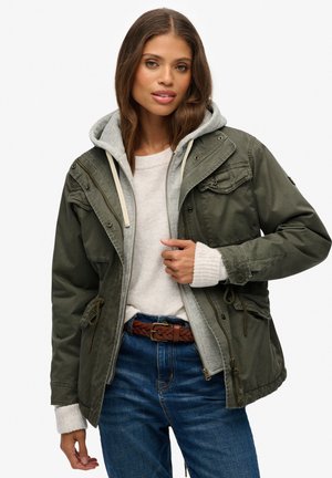 Olijfgroene jas met capuchon, voorzien van een ritssluiting en een koord, gelaagd over een lichtgrijze trui en blauwe jeans, met een gevlochten bruine riem.