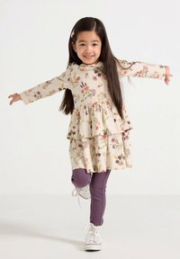 Crèmekleurige jurk met bloemmotief en gelaagde ruches, gecombineerd met geribbelde paarse legging en witte sneakers, met lange mouwen en een ronde halslijn.