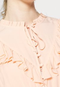 Persikka paita, jossa on röyhelökaulus, nappisuljenta ja herkkä solmukoriste. Pehmeä kangasrunko, jossa on kerroksellisia röyhelöitä edessä.