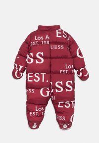 Raudonas pūkuotas snowsuit'as, aptraukta didžiuliu baltu "GUESS" prekės ženklu, su siuvinėtomis pagalvėlėmis, elastingais rankogaliais ir aukšta apykakle šilumai.