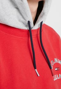 Sweatshirt rouge avec capuche grise et cordons bleu marine, montrant un texte partiel et un logo sur la poitrine près de l'encolure.