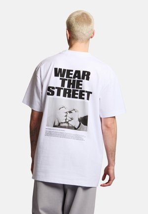 Osoba z krótkimi blond włosami nosząca białą, oversize'ową koszulkę z dużym czarnym napisem „WEAR THE STREET” oraz zdjęcie dwóch dłoni składających różowe przyrzeczenie.