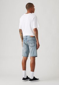 Tricou alb cu guler rotund, pantaloni scurți din denim bleu deschis și adidași negri cu tălpi albe. Prezintă tatuaje vizibile și un design simplu.