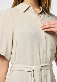 Abito camicia in lino beige con maniche corte, colletto, bottoni frontali e una cintura annodata in vita. Presenta una finitura testurizzata e una tasca sul petto.