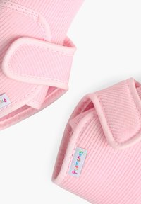 Des bottines en velours côtelé rose présentent une texture douce, une forme arrondie et des bandes Velcro. Une petite étiquette de marque affiche des lettres colorées.