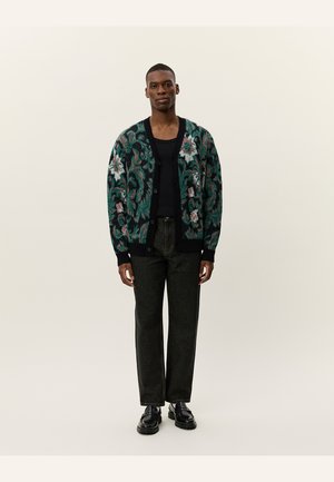 Knitted cardigan with a dark green base, floral patterns in pink and white, long sleeves, and front buttons; paired with dark pants and shoes.