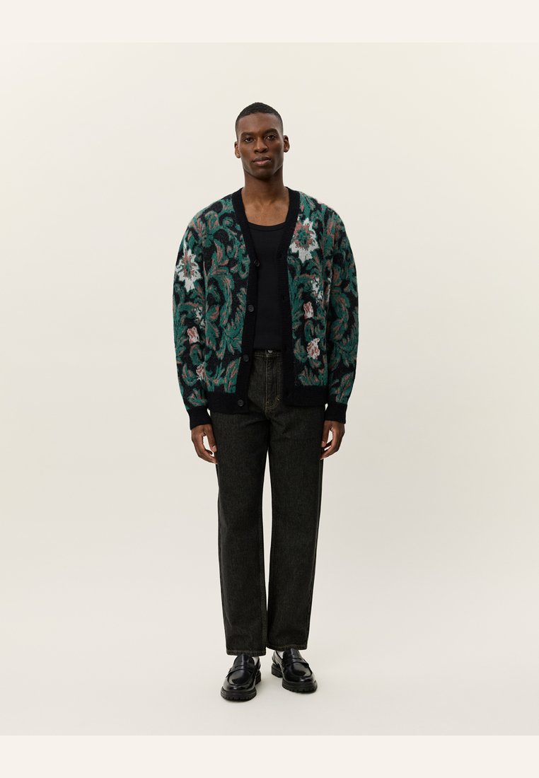 Cardigan tricoté avec une base vert foncé, des motifs floraux en rose et blanc, des manches longues et des boutons à l'avant ; associé à un pantalon foncé et des chaussures.