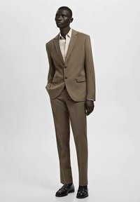 Hellbrauner Maßanzug mit einreihigem Sakko, Stegkragen und zwei Vordertaschen, kombiniert mit einem hellbeigen Hemd und schwarzen Schuhen.