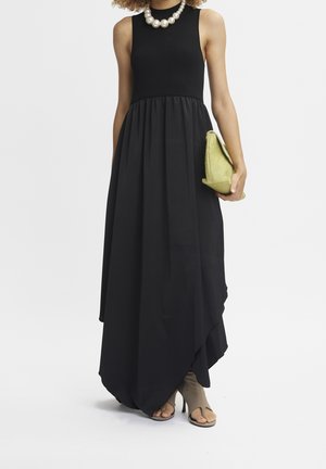 Robe maxi noire sans manches avec taille froncée, associée à un grand collier de perles, des talons ouverts couleur sable et une pochette vert clair.