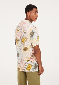 Camisa de manga corta en tejido ligero con un patrón geométrico colorido que presenta acentos en dorado, rosa, azul y verde. Cuello estándar y corte relajado.