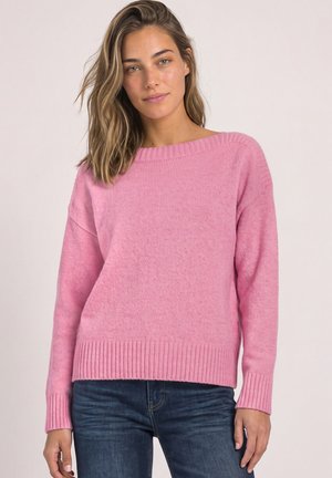 Femme aux cheveux mi-longs portant un pull en maille rose et un jean bleu, posant devant un fond clair uni.