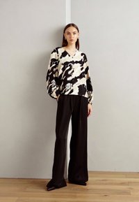 Blusa astratta in bianco e nero con maniche lunghe, abbinata a pantaloni neri larghi, su un pavimento in legno contro una parete neutra.
