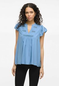 Blue short-sleeve blouse with a V-neck, textured fabric, decorative stitching, and a loose fit, worn with black pants.