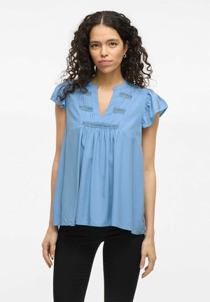 Blouse bleue à manches courtes avec un col en V, tissu texturé, coutures décoratives et coupe ample, portée avec un pantalon noir.