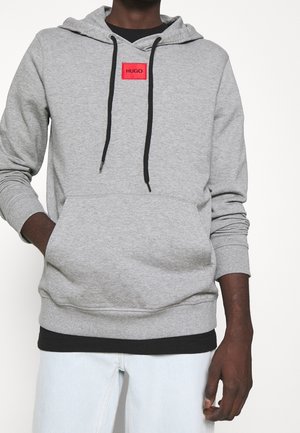 Personne portant un sweat à capuche gris clair avec des cordons noirs et un patch rouge étiqueté "HUGO,", accompagné d'un jean bleu clair et d'un sous-vêtement noir.