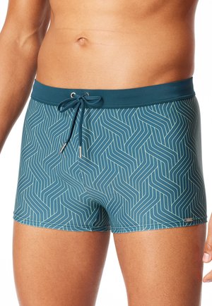 Männlicher Oberkörper mit türkisfarbenen Badehosen mit hellen geometrischen Linien und einem gebundenen Kordelzug in der Taille vor weißem Hintergrund.