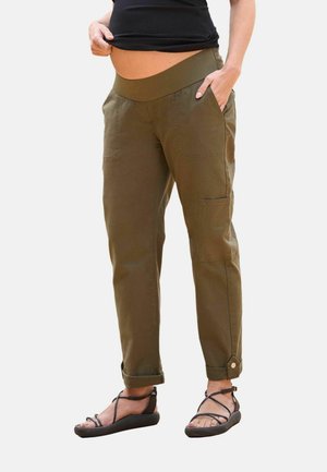 Woman wearing olive green maternity cargo pants with cuffed ankles and a black top, standing with one hand in pocket and one lifting shirt.