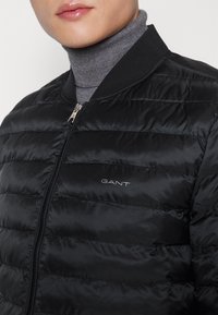 Svart puffjacka med horisontell baffeldesign, dragkedjeslutning och GANT-logotyp som visas på vänster sida. Hög krage, mjuk textur.