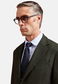 Giacca da completo verde a spina di pesce con risvolti, camicia azzurra chiara, cravatta blu navy testurizzata e occhiali neri rotondi. Capelli corti e pettinati all'indietro.