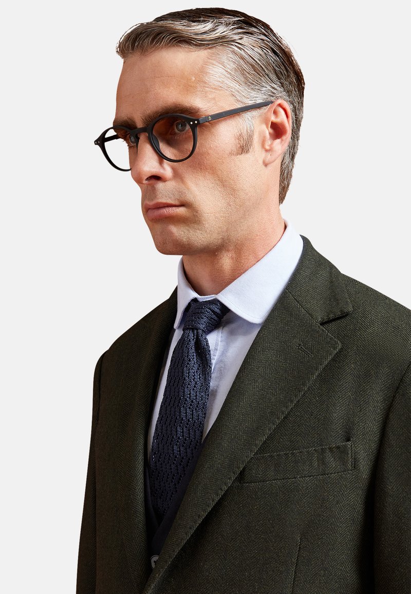 Giacca da completo verde a spina di pesce con risvolti, camicia azzurra chiara, cravatta blu navy testurizzata e occhiali neri rotondi. Capelli corti e pettinati all'indietro.
