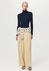 Vrouw in een marineblauwe turtleneck, beige wijde broek, gestreepte trui om haar taille geknoopt, en zwarte open teen sandals, staand tegen een effen achtergrond.