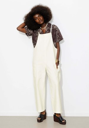Femme aux grands cheveux bouclés portant une salopette blanche, une chemise à motifs foncés, un collier épais et des sandales à plateforme noires, debout et posant.