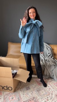 Svetlo modra jeans srajca z lokom na ovratniku, z zasukanim sprednjim delom na gumbe, dolgimi rokavi, kombinirana s črnimi hlačami. V bližini je kartonska škatla.