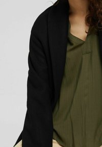 Blazer noir à l'avant ouvert avec une texture douce sur un blouse vert texturé présentant un motif rayé subtil et un décolleté en V.