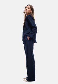 Marineblauer Nadelstreifenanzug mit einem taillierten Blazer und geraden Hosen. Weicher Stoff. Modell trägt hellfarbene Schuhe, die Hände in den Taschen.