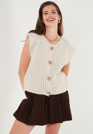 Young woman smiling wearing a cream knit sleeveless cardigan with gold buttons and a dark pleated skirt against a white background.