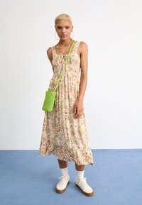 Floral maxi-jurk met pastelkleuren, gerimpelde buste en een ruchezoom. Gepaarde met een kleine groene gehaakte tas en witte sneakers.