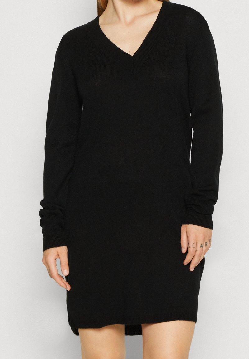 Femme portant une robe pull noire à manches longues avec un col en V, un tatouage sur la main droite et un teint clair, debout devant un fond uni.