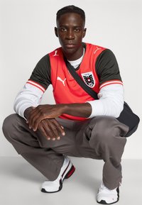 Piros és fekete Puma sportmez rövid ujjakkal, szürke katonai nadrág és fehér, piros részletekkel díszített sportcipők. Modell guggoló helyzetben.