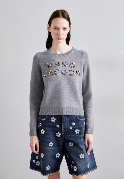 Harmaa neulepusero, jossa on "MARC JACOBS" -logo värikkäillä koristeilla, yhdistettynä sinisiin farkkushortseihin, joissa on valkoista kukkakirjontaa.