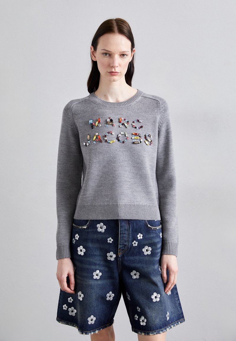 Suéter de punto gris con el logotipo "MARC JACOBS" en adornos coloridos, combinado con pantalones cortos de mezclilla azules que presentan bordados florales blancos.