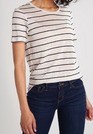 Jasnobeżowa koszulka z granatowymi poziomymi paskami, o okrągłym dekolcie, krótkich rękawach i luźnym kroju, zestawiona z ciemnoniebieskimi jeansami.