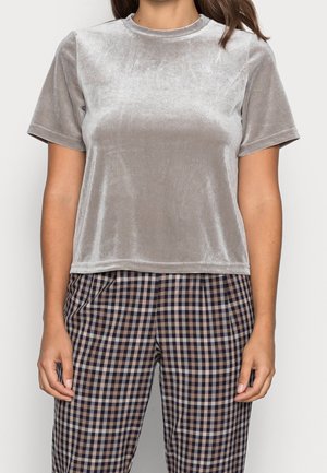 Vrouw draagt een lichtgrijs fluwelen shirt met korte mouwen en een hooggetailleerde, bruin-blauw geruite broek tegen een effen achtergrond.