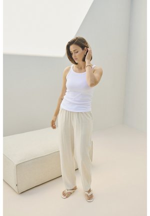 Un débardeur blanc associé à un pantalon beige clair ample et des sandales plates. L'arrière-plan présente des couleurs claires et des textures douces.