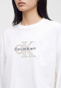 Γυναίκα που φοράει λευκό φούτερ με λογότυπο Calvin Klein σε ανοιχτό μπεζ και σκούρο μπλε στο στήθος, εμφανίζοντας τον λαιμό και το κάτω μέρος του προσώπου.