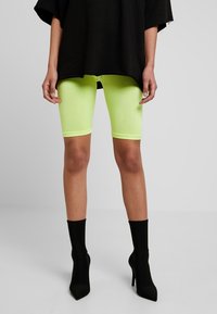 Shorts da bici verde neon in tessuto elasticizzato, abbinati a una maglia nera larga oversize e stivaletti neri con tacco alto.