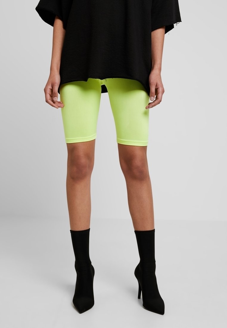 Shorts da bici verde neon in tessuto elasticizzato, abbinati a una maglia nera larga oversize e stivaletti neri con tacco alto.
