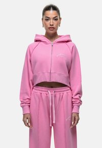 Felpa corta rosa con zip frontale, cappuccio e logo ricamato. Abbinata a pantaloni da tuta rosa coordinati con vita elasticizzata.